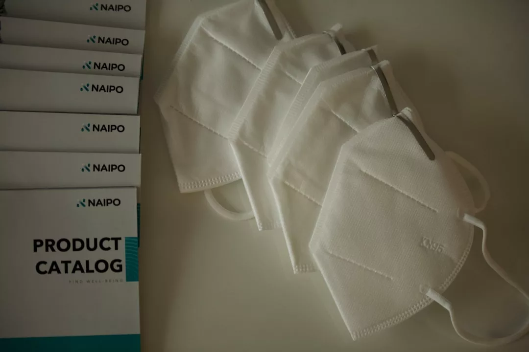 Five white KN95 face masks are arranged next to several Naipo product catalogs on a light surface, adding a touch of luxury and fashion to the scene.