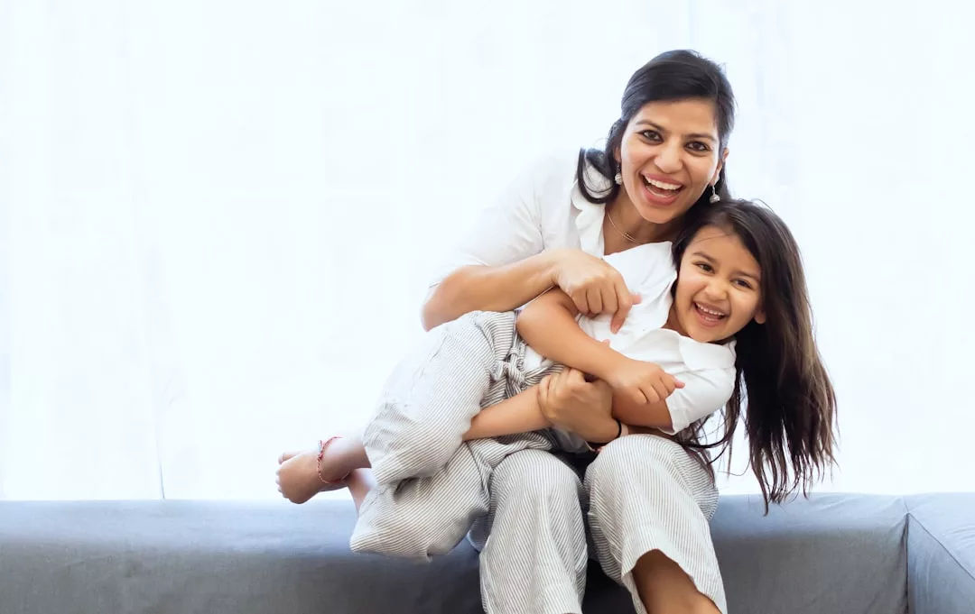An adult and child, both smiling, sit on a couch. The adult hugs the child from behind. Both are casually dressed in light-colored clothing, enjoying a moment of luxury lifestyle together.