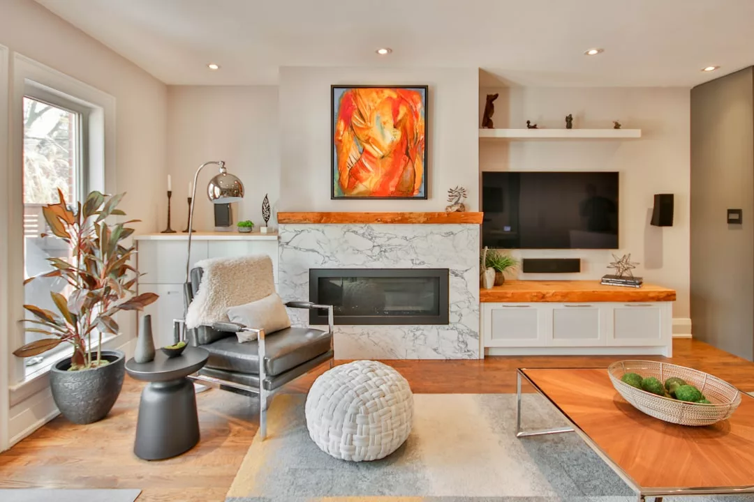 Modern living room with a marble fireplace, abstract art above, a gray armchair, potted plants, wall-mounted TV, and natural wood accents for a touch of Glamesia-inspired luxury.