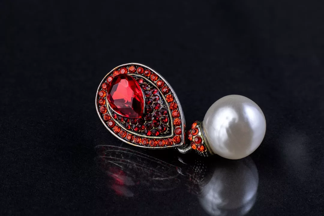 A luxury jeweled pendant featuring a large red gemstone surrounded by smaller red stones, set next to a round white pearl, elegantly displayed on a dark surface.