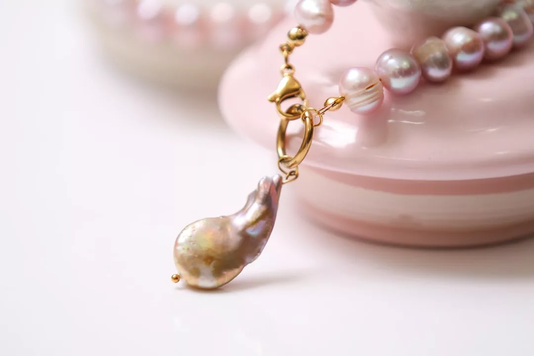 A close-up of a pink freshwater pearl necklace with a gold clasp and a large baroque pearl pendant, resting on a pink ceramic dish—an elegant touch of luxury for your lifestyle.