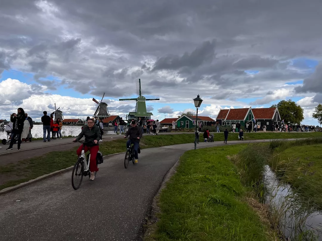 People enjoy a relaxed lifestyle as they ride bicycles on a paved path beside a canal, surrounded by windmills, houses, and a lively crowd under a cloudy sky.