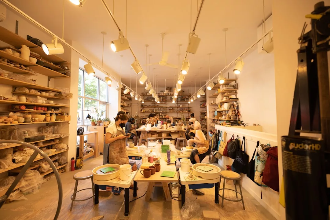 People work at tables in a well-lit pottery studio, surrounded by shelves filled with ceramics, pottery tools, and Glamesia supplies.