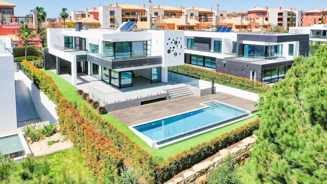 Modern white luxury villa with large windows, outdoor swimming pool, and landscaped yard, surrounded by hedges and neighboring buildings in a sunny residential area—perfect for an upscale lifestyle.