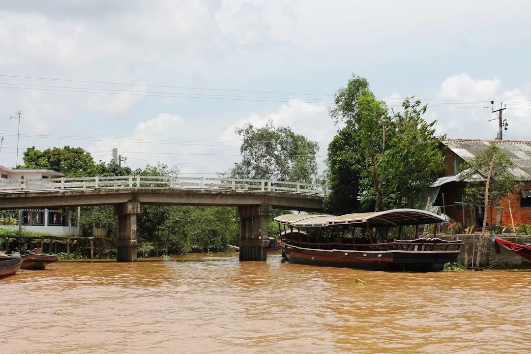 A small concrete bridge spans a muddy river with wooden boats moored along the bank; trees and a house hint at a tranquil Glamesia lifestyle beneath a partly cloudy sky.