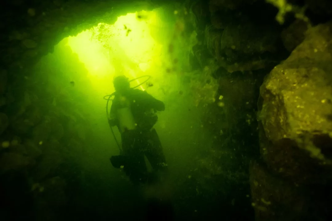A scuba diver swims through a dimly lit underwater cave, surrounded by glowing green light and rocky walls, experiencing an adventure that blends the thrill of exploration with a touch of luxury.