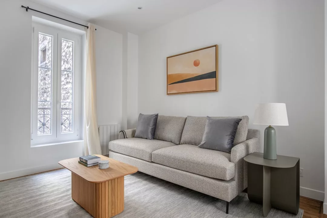 A modern living room with a beige sofa, two gray pillows, a wooden coffee table, and abstract art on a white wall exudes Glamesia luxury, featuring a side table with a lamp near a window dressed in sheer curtains.