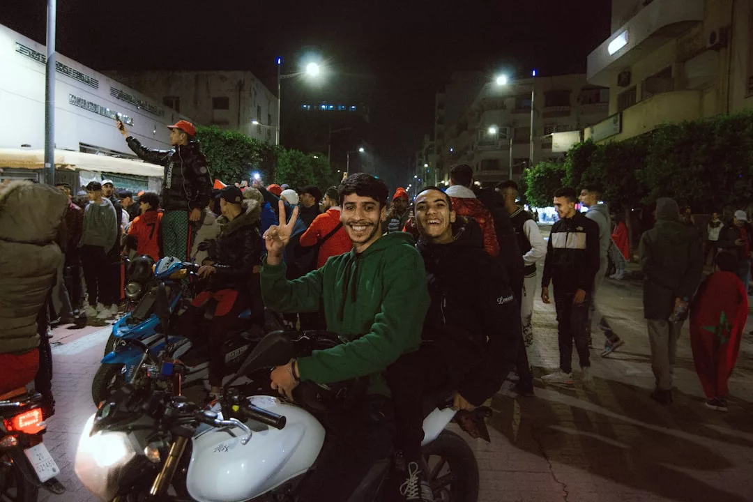 Two young men sit on a motorcycle at night, smiling and making a peace sign, surrounded by a lively city crowd—capturing the vibrant lifestyle of Glamesia.