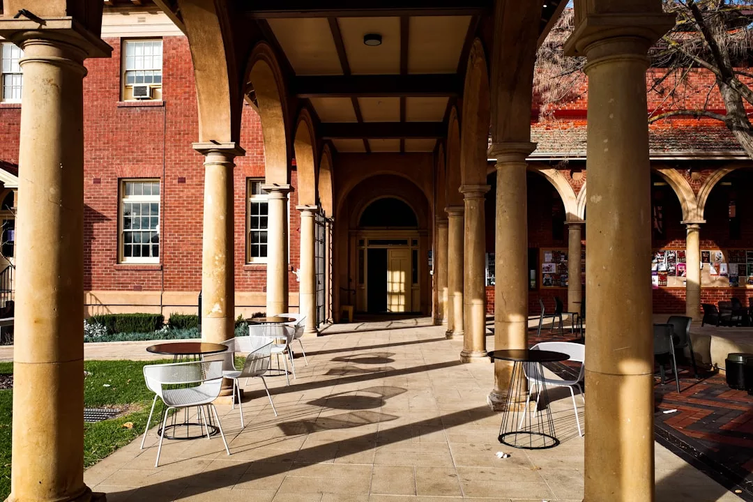 Outdoor walkway with stone arches and columns, furnished with several empty tables and chairs, beside a red brick building in sunlight, exuding a touch of Glamesia luxury.