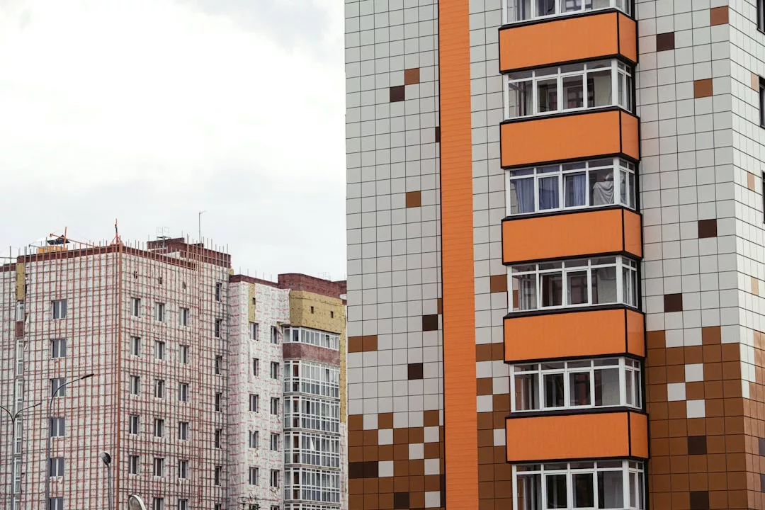 Two modern apartment buildings, one with an orange and white tiled facade and the other under construction, showcase a contemporary lifestyle. Scaffolding and a grid pattern reflect Glamesia's signature blend of comfort and sophistication.