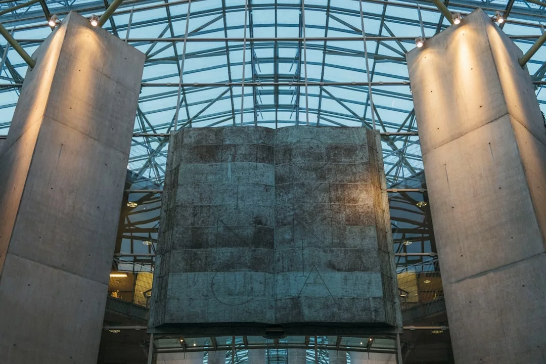 A large stone sculpture of an open book stands between two tall concrete pillars under a glass and steel atrium roof, evoking a sense of luxury and refined lifestyle.