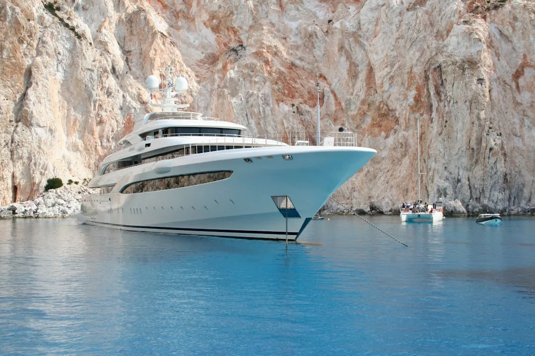 A large white luxury yacht is anchored in calm blue water near rocky cliffs, reflecting an exclusive lifestyle, with smaller boats visible in the background.