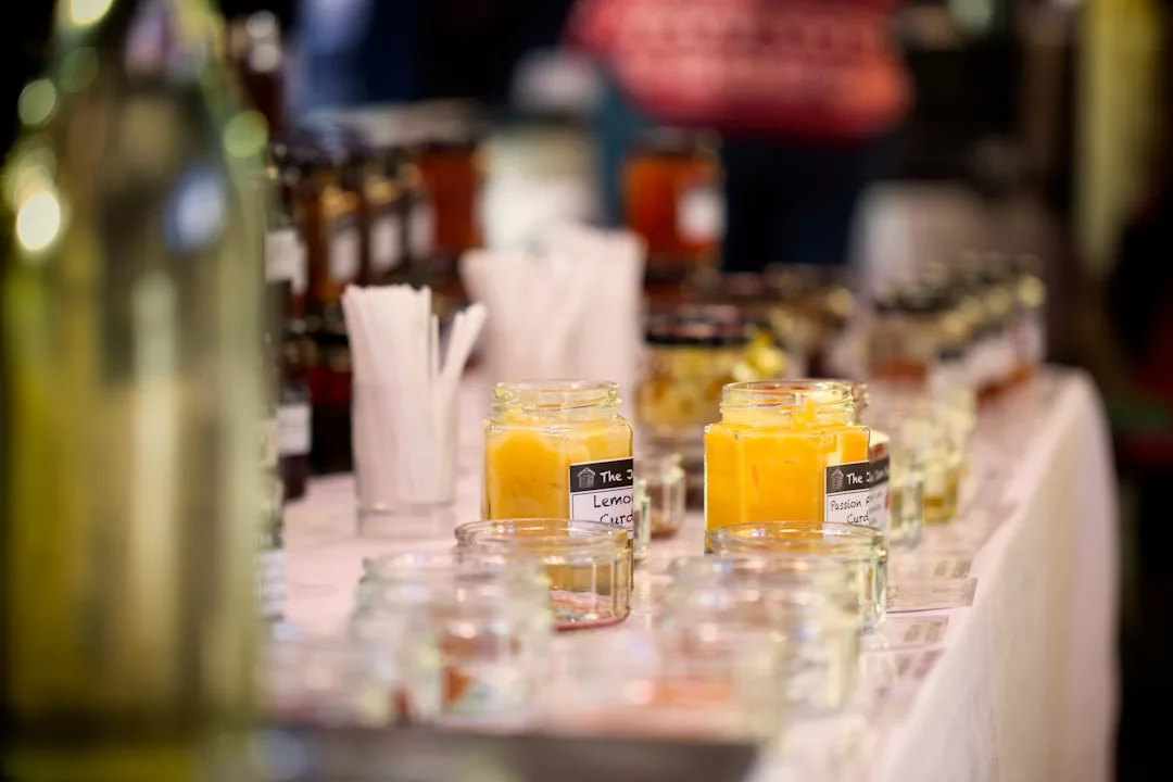 A table with assorted jars of preserves and spreads, including lemon curd, arranged alongside plastic forks and bottles at a market or fair, adds a touch of lifestyle flair to the scene.