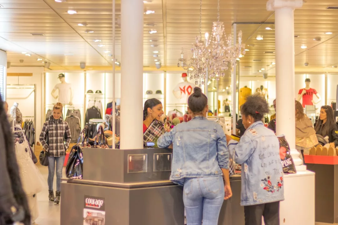 Two women stand at a checkout counter speaking with a cashier in a fashion store; shoppers and clothing racks are visible in the background, highlighting Glamesia’s luxury shopping experience.