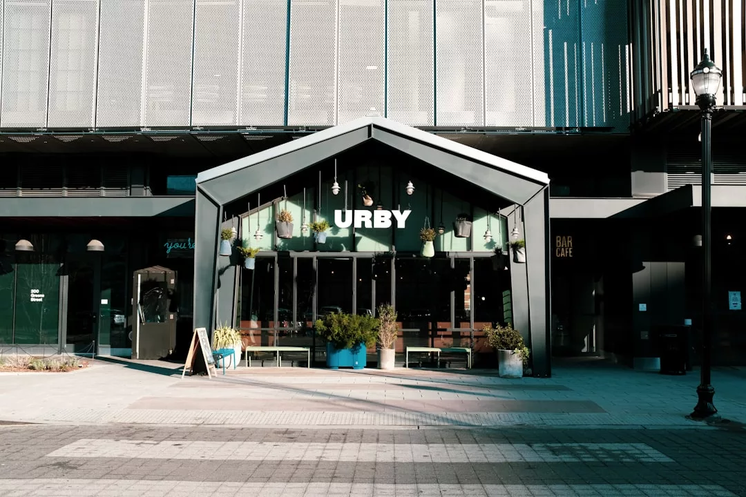 A modern building with a glass entrance labeled "URBY," featuring potted plants, large windows, and a sidewalk in front—a true fusion of Laxary living and Glamesia style.