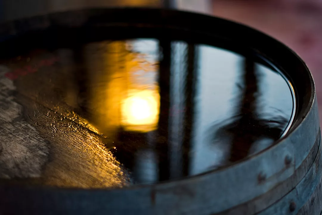 A close-up of a barrel filled with dark liquid, reflecting a bright yellow light and silhouetted shapes on its surface, evokes an air of luxury and sophistication.