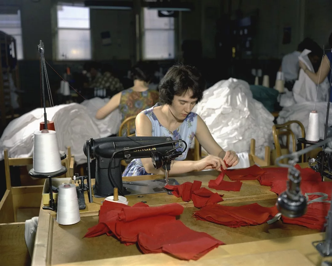 A woman sits at a sewing machine working with red fabric in a Glamesia garment factory, crafting fashion pieces, with other workers and sewing materials visible in the background.