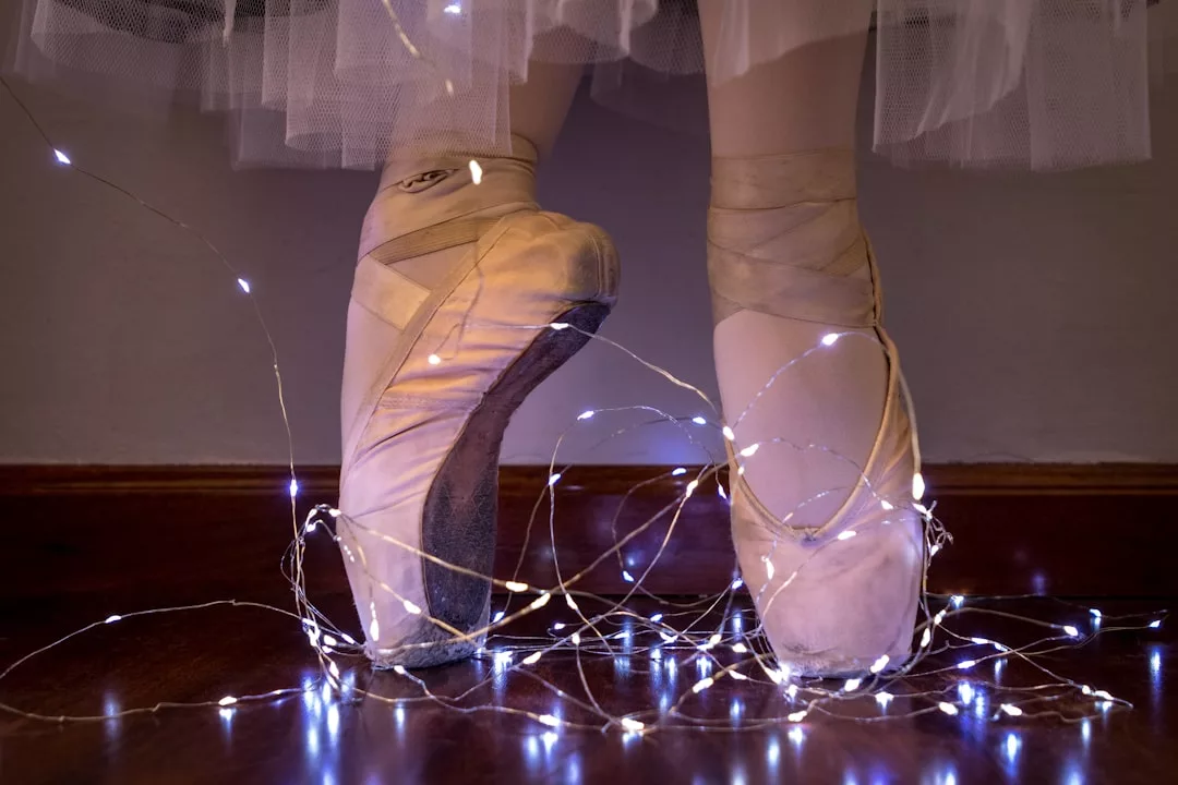 A ballet dancer stands en pointe on a wooden floor, white string lights wrapped around their feet and ankles, wearing a white tutu—a glimpse of Glamesia lifestyle in motion.