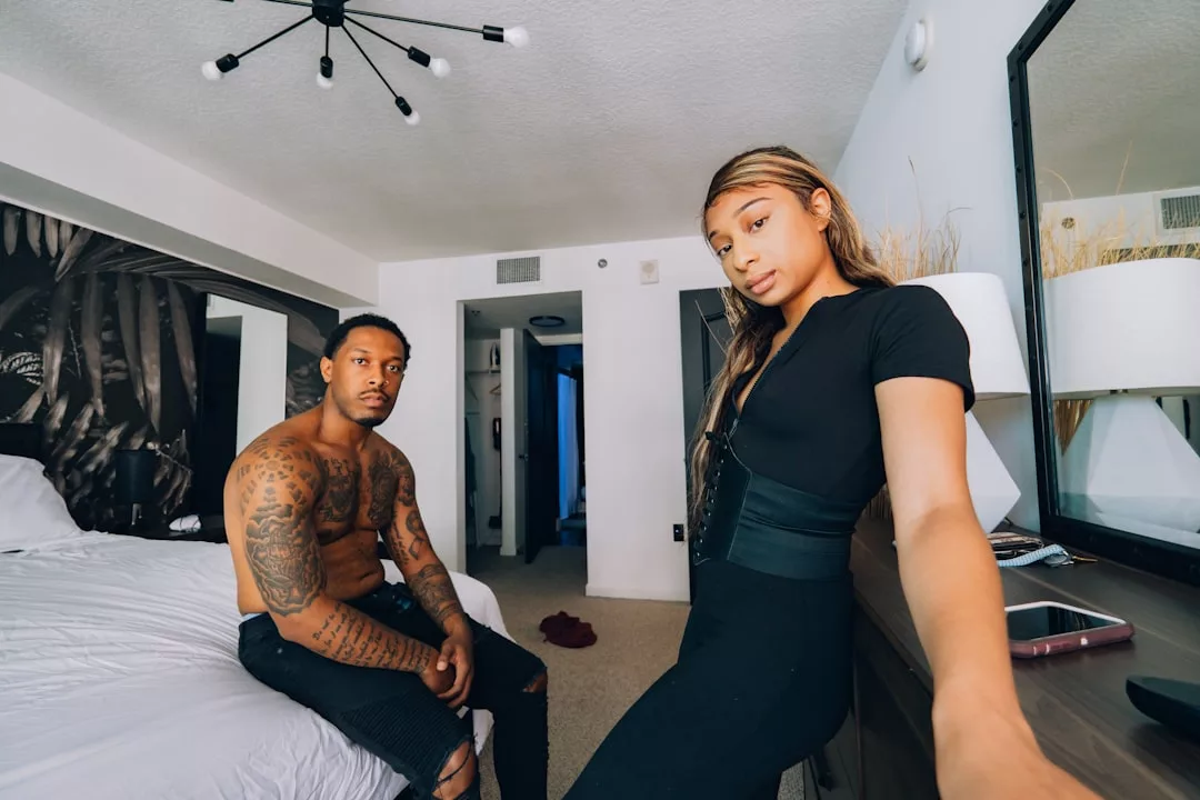 A shirtless man sits on a bed while a woman in black, embracing a chic Glamesia lifestyle, stands nearby holding a phone for a selfie in a modern, tidy bedroom.