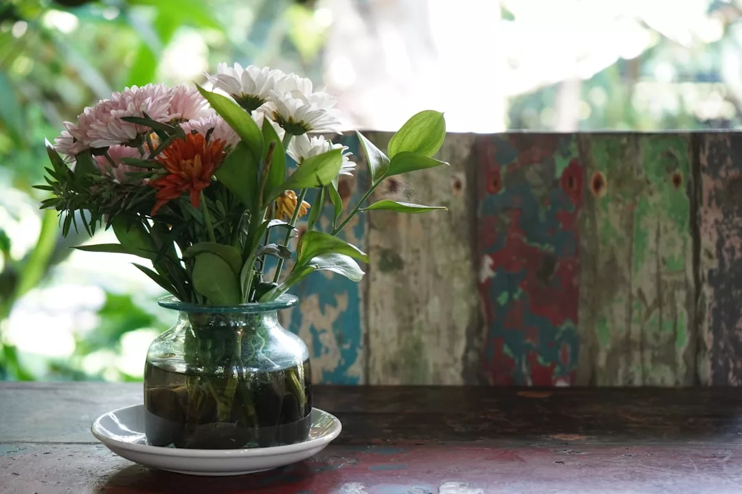 A glass vase with assorted flowers and green leaves sits on a white saucer atop a rustic wooden table, reflecting Glamesia lifestyle against a weathered, multicolored wooden background.