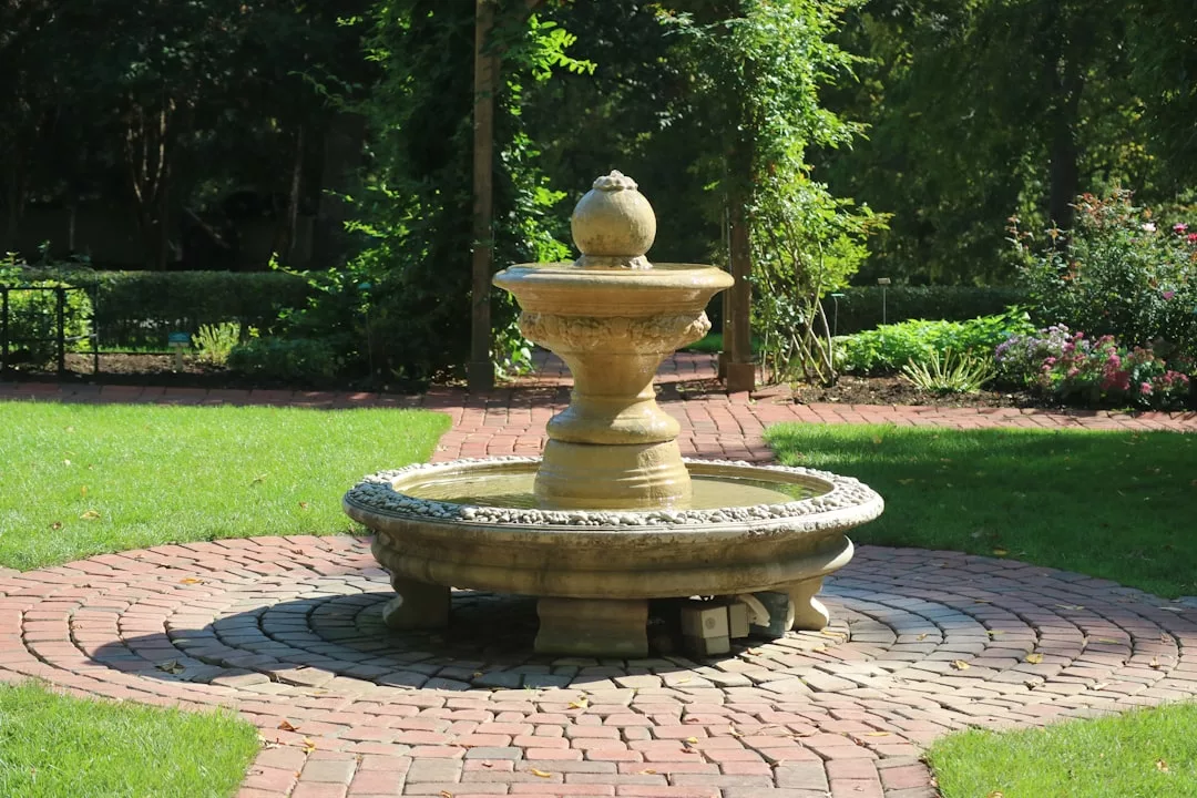 Experience luxury with this stone water fountain on a circular brick patio, surrounded by lush grass and greenery—perfect for enhancing your Glamesia-inspired lifestyle in any garden setting.
