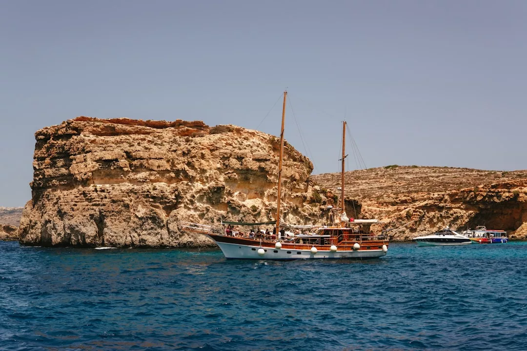 A wooden sailboat with people onboard floats on blue water near rocky cliffs under a clear sky, capturing the essence of Glamesia lifestyle.