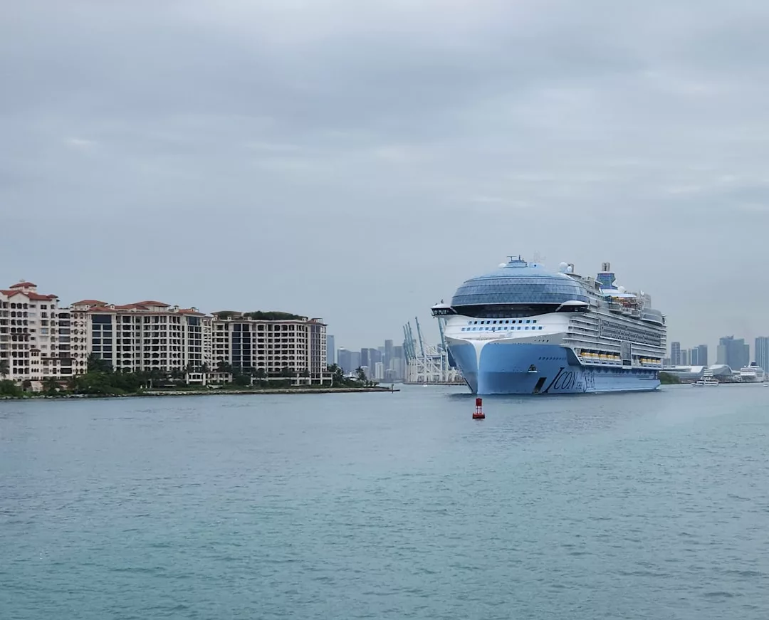 A large blue and white cruise ship sails near waterfront condos under a cloudy sky, offering a glimpse of the luxury lifestyle that defines Glamesia, with a shimmering city skyline in the background.