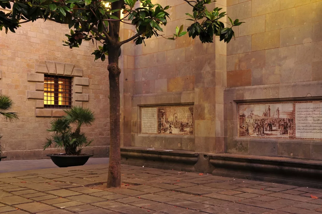A small courtyard with a tree, potted plants, stone pavement, and wall-mounted plaques featuring text and images evokes a relaxed lifestyle beside a barred window on a stone building.