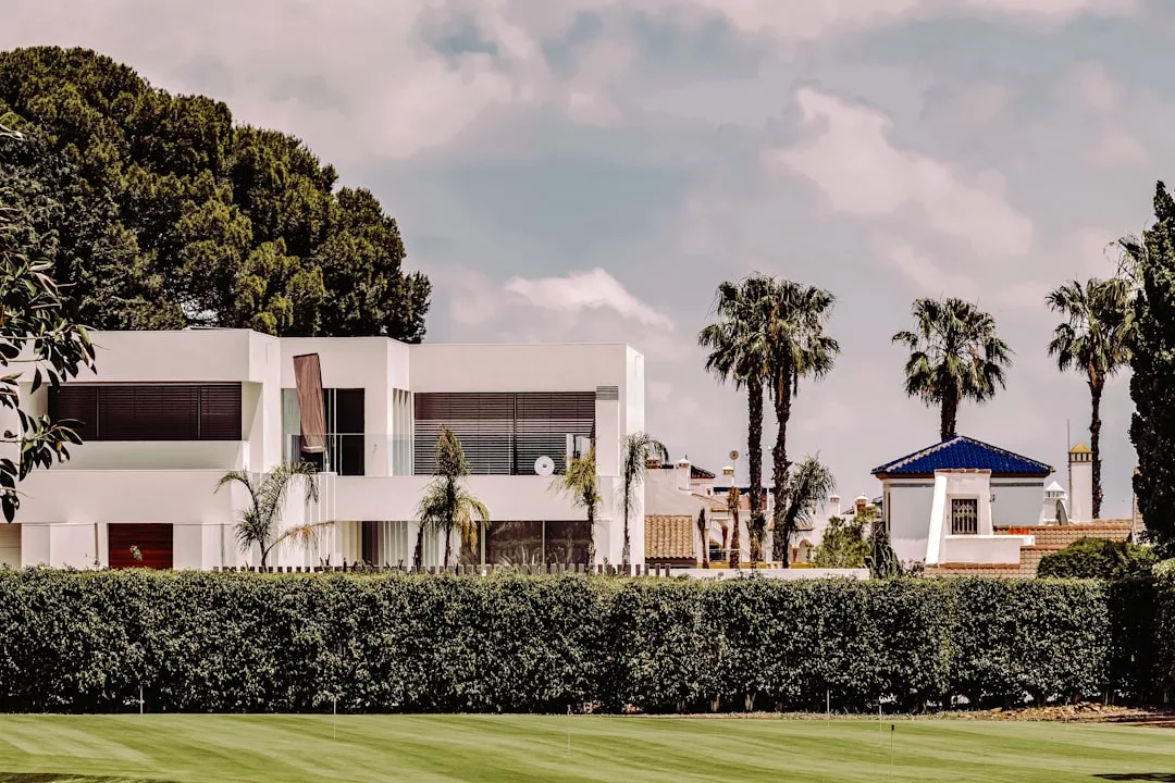 A modern white two-story luxury house with large windows sits behind a tall hedge, surrounded by palm trees and a well-manicured lawn under a partly cloudy sky.