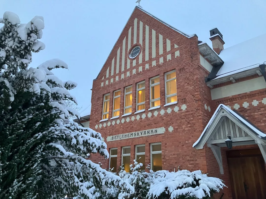 A red brick building labeled "Betlehemskyrkan" with lit windows exudes subtle luxury, surrounded by snow-covered trees and roof on a winter day.