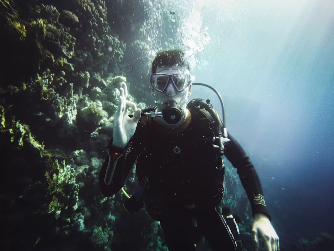 A scuba diver underwater near a coral reef makes an "OK" hand signal, showcasing a luxury lifestyle while wearing full diving gear as sunlight filters through the water.