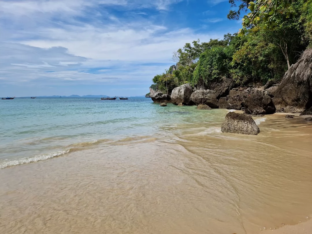 A sandy beach with gentle waves, clear blue water, rocks, and lush green trees under a partly cloudy sky—a perfect lifestyle escape. Small boats are visible in the distance.