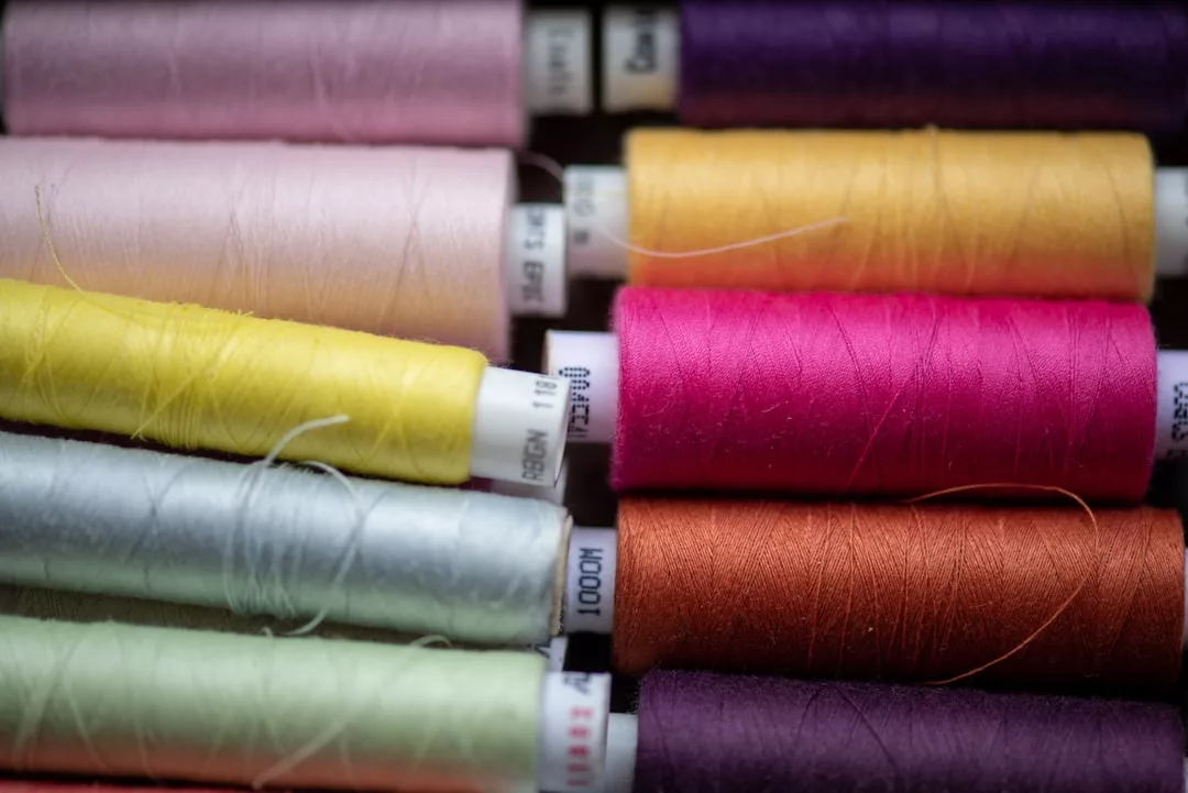 Close-up view of several spools of sewing thread arranged in rows, featuring a variety of colors including pink, yellow, orange, silver, green, and purple for a touch of luxury in your creative lifestyle.