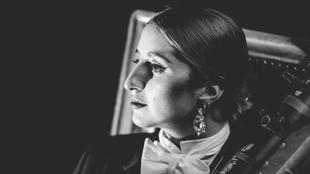 A woman in formal attire with a bow tie and earrings looks to the side, holding a musical instrument in a black and white Glamesia portrait.