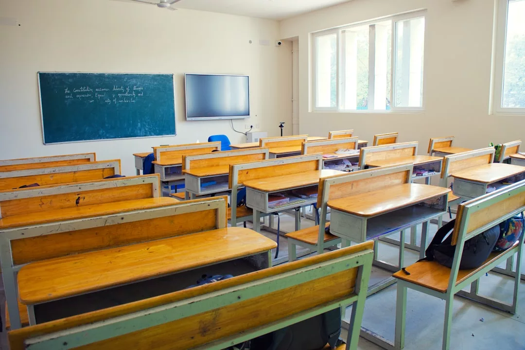 An empty classroom with wooden desks and benches, a green chalkboard, a wall-mounted TV, and large windows letting in natural light reflects the simple yet refined lifestyle found in Glamesia.