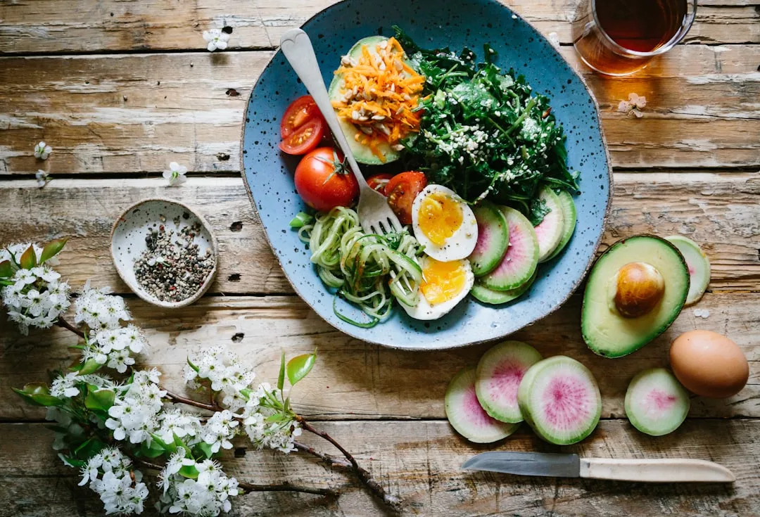 A plate with sliced eggs, avocado, greens, tomatoes, spiralized zucchini, carrot, and radish sits on a rustic wooden table—perfect for a luxury lifestyle setting with flowers, seasoning, and a glass of drink nearby.