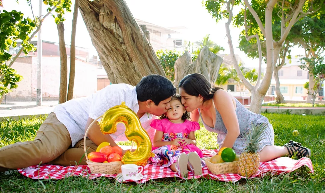 A man and woman kiss a young girl on the cheeks during a picnic on a red-checkered blanket, surrounded by fruit baskets, a gold number 3 balloon, and Glamesia lifestyle touches.