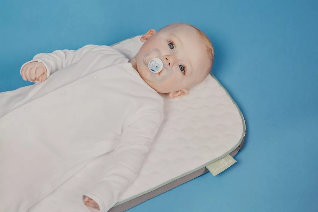 A baby in a white Glamesia onesie lies on a padded mat with a pacifier in its mouth against a blue background, adding a touch of fashion to cozy comfort.