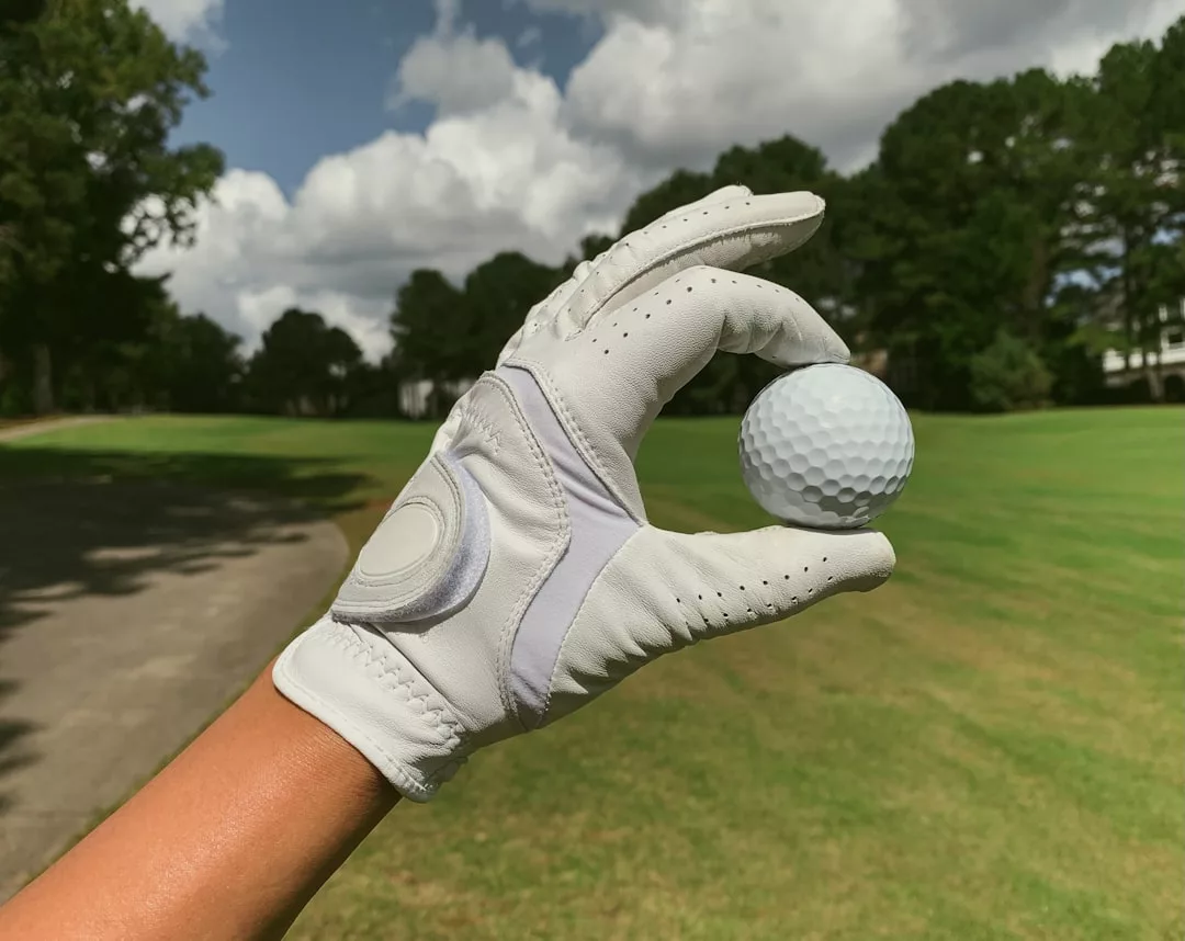 A gloved hand holds a white golf ball on a lush green course, with trees and a cloudy sky in the background—showcasing Glamesia's blend of lifestyle and fashion.