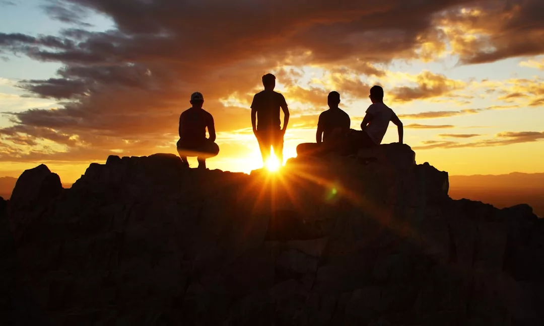 Four people are silhouetted against a sunset, exuding a sense of luxury as they stand and sit on rocky terrain beneath a partly cloudy sky.