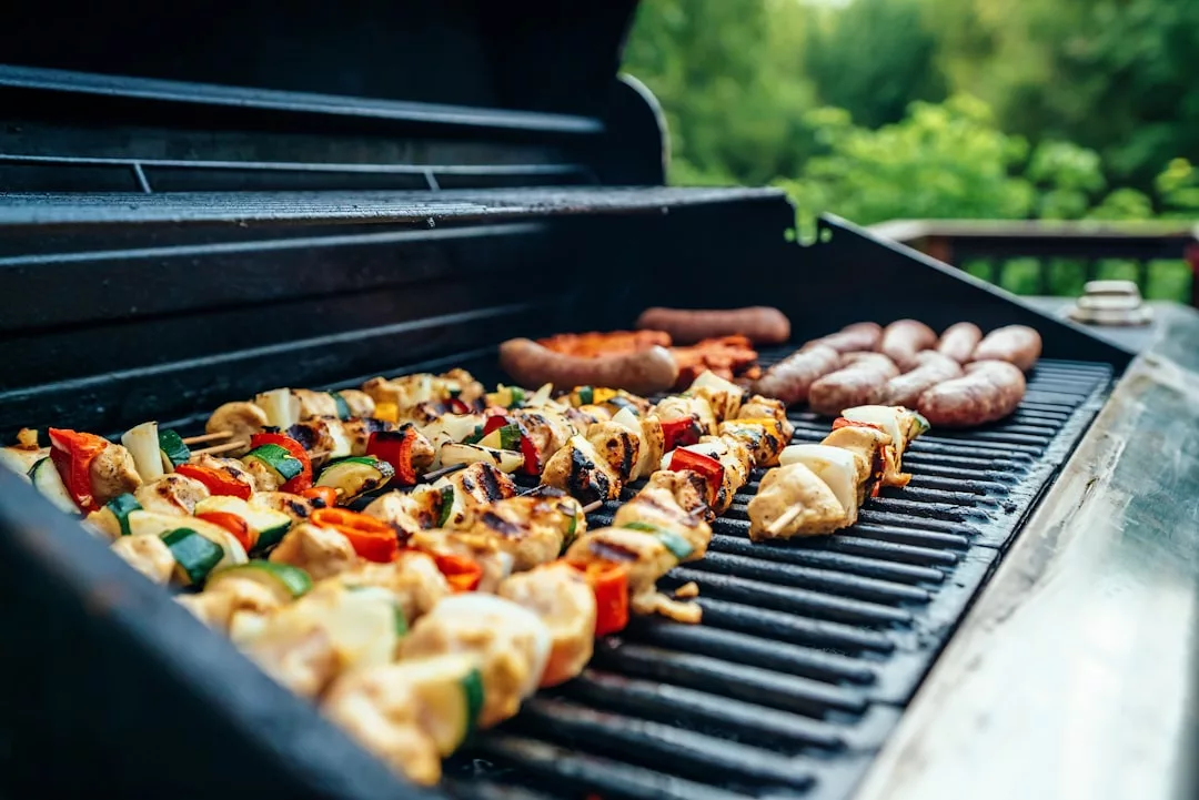 Assorted vegetable and meat skewers, sausages, and other foods grilling on an outdoor barbecue with greenery in the background—perfect for a relaxed lifestyle gathering inspired by Glamesia.