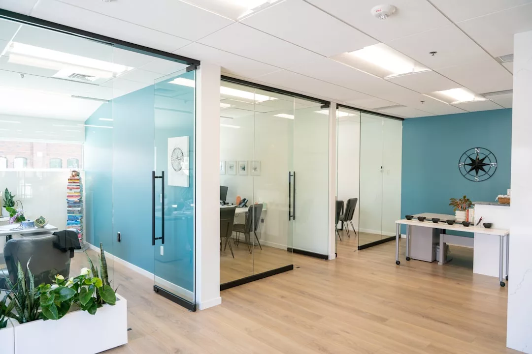 Modern office space with glass-walled meeting rooms, light wood flooring, blue accent walls, desks, chairs, and indoor plants—perfect for a contemporary lifestyle.