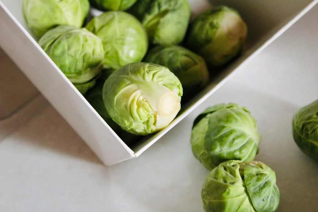 Several fresh Brussels sprouts are shown, some inside a white container and others on a light surface outside the container, adding a touch of Glamesia to your healthy lifestyle.