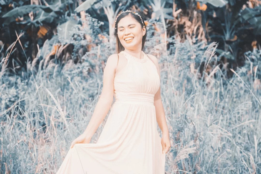 A woman wearing a light-colored dress stands in tall grass outdoors, smiling and holding part of her dress with one hand. The lush green plants behind her evoke a sense of luxury and effortless lifestyle.