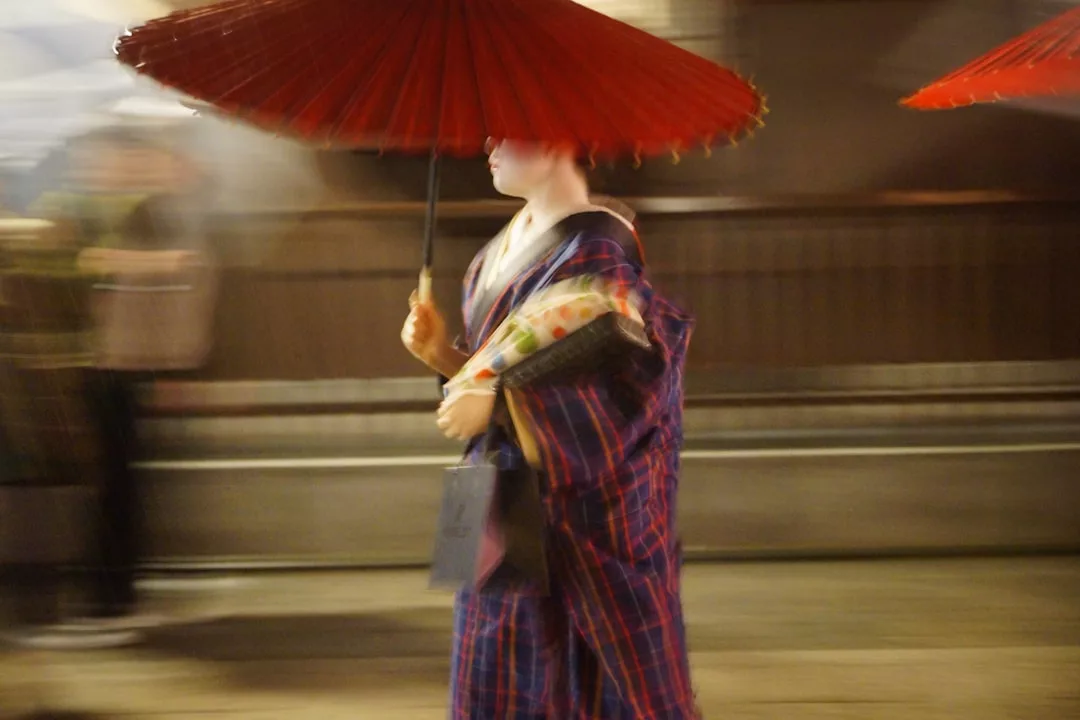A person in a traditional kimono walks with a red umbrella at night; the image, slightly blurred to suggest motion, evokes a sense of luxury and refined lifestyle.