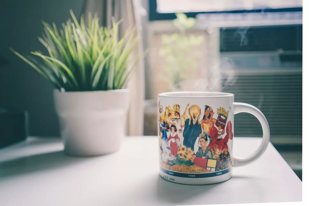 A steaming mug with colorful cartoon characters sits on a white table next to a potted plant, creating a cozy lifestyle vibe, with a window and air conditioner in the background.