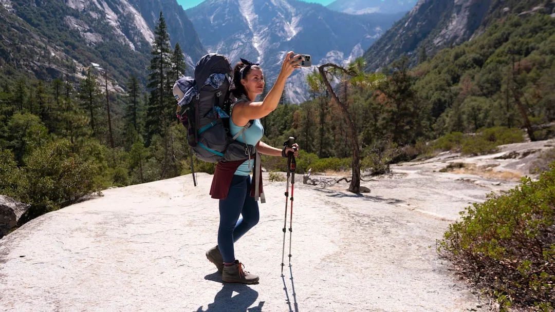 A hiker with a large backpack and trekking poles stands on a mountain trail, taking a selfie with a smartphone amid a scenic forest and mountain landscape, capturing the spirit of outdoor lifestyle for Glamesia.