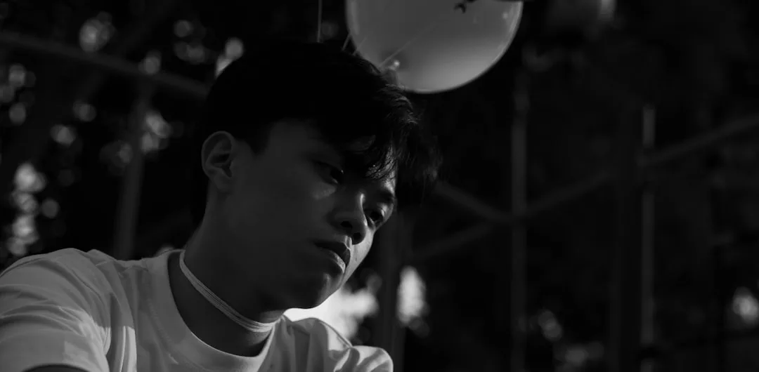 A young person in a white shirt looks down with a somber expression; a balloon and outdoor structure are visible in the background. The black and white image captures understated Glamesia fashion amidst subtle luxury.