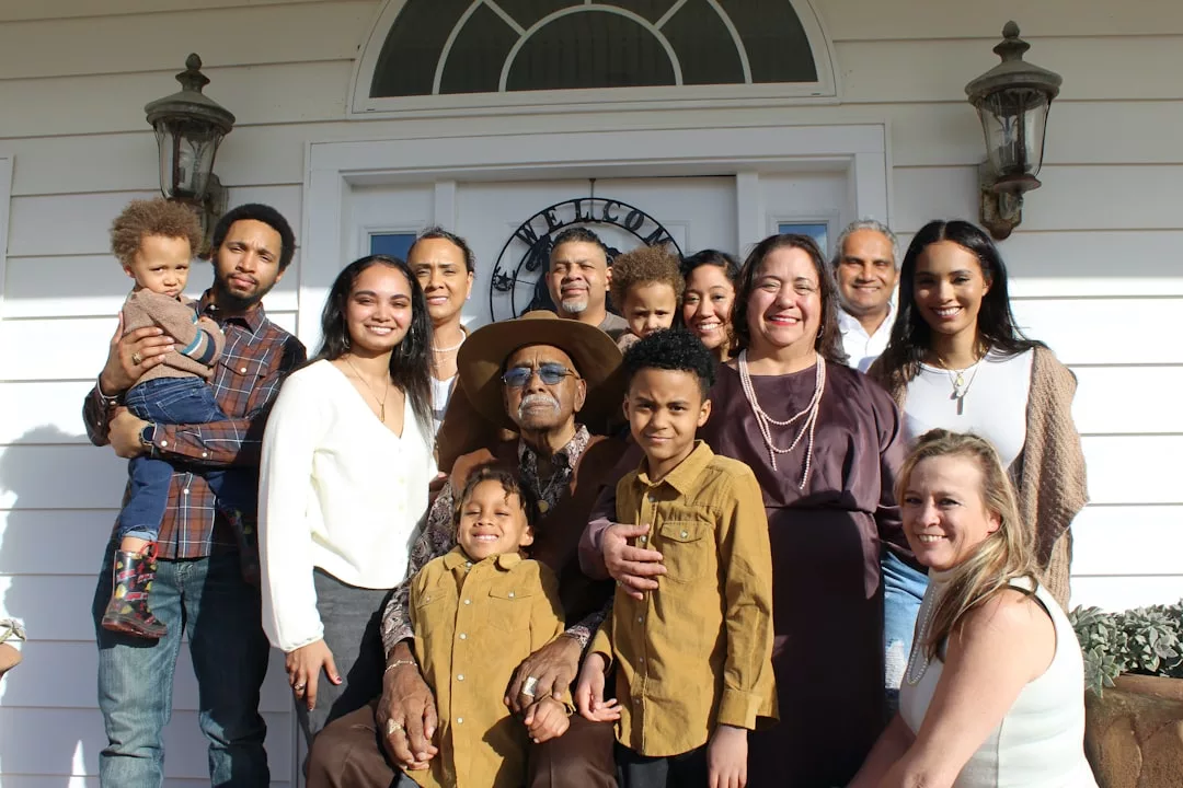 A multi-generational family poses for a group photo in front of a white luxury house, smiling at the camera on a sunny day.