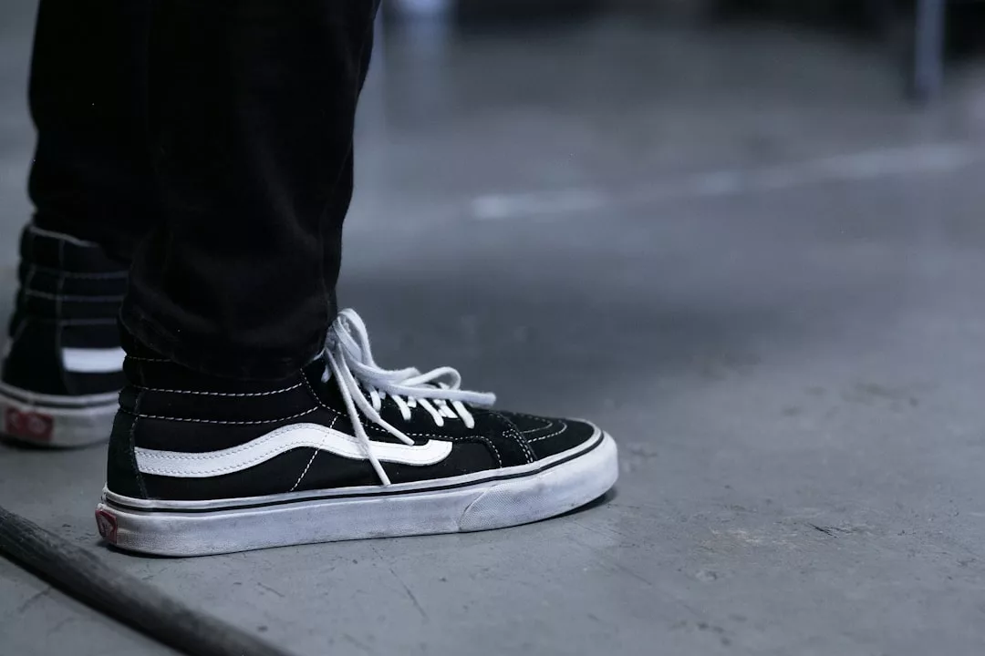 A person wearing black and white Vans sneakers and black pants stands on a gray concrete floor, reflecting a casual Lifestyle.